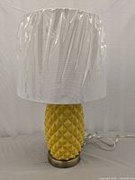 Front side view of the yellow pineapple ceramic lamp base with white fabric cylindrical lampshade covered in plastic.