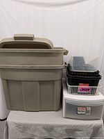 Two large beige Rubbermaid storage totes with lids stacked, smaller black, clear, and grey plastic baskets and containers beside them on a table.
