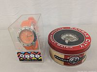 Wets orange wristwatch in clear plastic box and Kansas City Railroad pocket watch in round tin box with lid featuring railroad design.