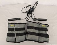 Photo of black skipping rope with plastic handles and two gray fabric ankle weights with black straps and metal rings laid out on a white surface.