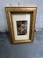 Photo of antique framed floral art print against a concrete background, showing the gold ornate frame and matting around the print.