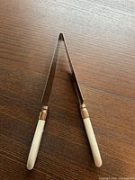 Top-down view of the cake slicer with cream-colored handles and gold inlay near the blade junction, blade arranged in a V shape.