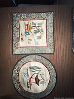 Photo of two Chinese silk tapestries, one square and one round, laid flat on a wooden surface showing hand-stitched temple and landscape scenes with patterned borders.