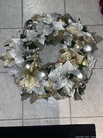 Top-down view of the wreath placed on a tiled floor showing silver and gold poinsettia flowers, green pine accents, and various silver and gold ornament balls.