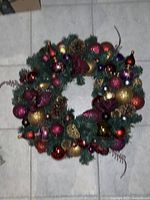 Full view of the custom handmade Christmas wreath placed on a tiled floor showing artificial pine foliage, pinecones, and multi-colored shiny baubles including red, gold, purple, and pink.
