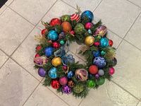 Top down view of round multi-coloured ornament Christmas wreath on tiled floor showing various baubles and green pine needles.