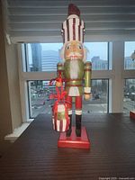 Front view of the green sparkly suited nutcracker on a red base, holding a gift bag with red ribbon, with cityscape background.
