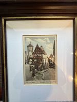 Framed signed and titled etching showing street scene with half-timbered architecture and clock tower.