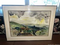 Framed watercolor landscape painting with hills, trees and dramatic sky with sun rays breaking through clouds.