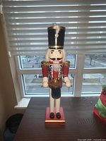 Front view of large classic nutcracker figure on a table by a window showing red jacket, white pants, and tall black hat.