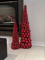 Photo of two tall red Christmas ornament ball trees of different sizes placed on a tiled floor near a fireplace.