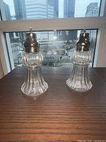Pair of antique glass salt and pepper shakers with silver tops shown on a wooden table with city background.