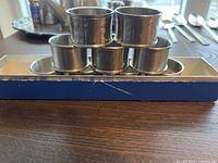 Front view of six polished silver napkin rings stacked in a pyramid on top of a blue and silver rectangular box. Surface scratches visible on box sides.