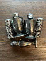 Four antique silver salt and pepper shakers on wooden surface, two solid silver and two with silver lattice design, showing tarnish and use typical for antique items.