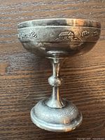 Full side view of the engraved silver wine cup showing grapevine and vineyard house motifs around the bowl and base on a wooden surface.