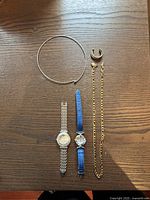 Photo of the lot showing two wristwatches (one silver-tone metal band, one blue leather strap), a gold-tone chain necklace, a silver-tone choker necklace, and a gold-tone ring shaped like a horseshoe on a wooden surface.
