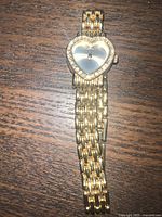 Full view of the gold-tone heart-shaped wristwatch on a wooden surface, showing the watch face and linked bracelet band.