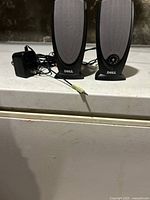 Two Dell computer speakers and an AC adapter placed on a white surface with a stone wall background. Speakers have gray grills and black stands, with visible control knobs on one speaker.