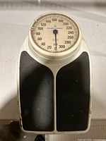 Top view of a Health o meter analog bathroom scale showing the round dial with weight markings up to 325 lbs. The scale platform has black footpads and a mainly white plastic casing with signs of wear and rust.