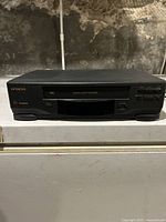 Front view of black Hitachi FX600 VHS player on white surface against a textured wall, showing front tape slot and control buttons.