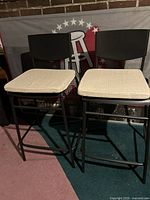 Two black metal counter stools with beige cushions shown from the front against a backdrop