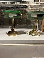 Pair of vintage bankers lamps showing the front view of green glass shades and brass bases with light bulbs and cords, placed on a white surface in a basement or garage.