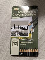 Front of green metal tin case showing printed label with charcoal pencils and landscape art.