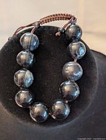 Front view of black obsidian bracelet on black display with large and small beads and brown braided adjustable cord.