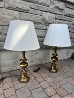 Two brass lamps with white shades placed on a stone pavement in front of a stone wall. Lamps are lit by outdoor light, showing polished brass bases and clean shades.
