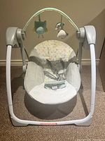 Front view of the baby swing showing cushioned seat, safety harness, and toy bar with hanging plush toys.