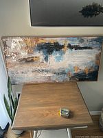 Framed abstract canvas art leaning against a wall next to a wooden table and plant.