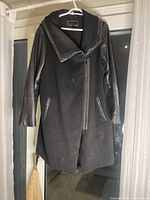 Front view of black women's coat hanging, showing leather sleeves and wool body, zipper closure, minor lint and dust visible.