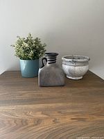 Side view of the faux plant pot in blue ribbed ceramic, a gray and navy blue square ceramic vessel, and white and gray marbled ceramic plant pot, all placed on a wooden surface against a plain wall.