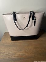Front view of the Kate Spade tote bag showing beige and black two-tone leather exterior, with black peeling handles and a gold logo plaque.