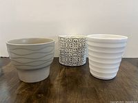 Three plant pots arranged in a row on a wooden surface against a plain background.