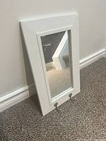 Photo showing the white framed mirror with two hooks leaning against a wall on carpet.