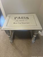 Top view of a small rectangular wooden table with light gray painted surface and decorative black print 'PARIS No. 9 Rue Le Verrier'.