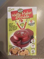 Front box image showing the red Grinch mini waffle and griddle maker and waffles with character shapes.