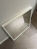 Top-down angled photo showing the white framed square mirror resting against a wall on carpeted floor.