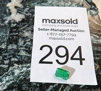 Emerald gemstone on auction card with lot number 294, showing size and color contrast against a dark marble surface.