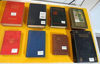 Eight antique hardcover books laid flat on yellow surface, showing spines and covers with dates and titles visible.