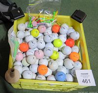 Yellow crate filled with around 100 golf balls of various colors and brands, some pink, orange, yellow, blue, mostly white. Small tan cone-shaped holder also visible.