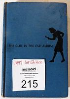 Front cover of the book showing blue hardcover with black silhouette and title The Clue in the Old Album plus the auction label indicating 1947 1st Edition.