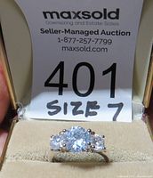 Close-up photo of the ring from the front showing three clear gemstones set in a silver band, inside a display box with a size 7 tag.