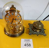 Photo showing vintage 400-day clock under glass dome and ornate brass inkwell side by side on yellow surface.