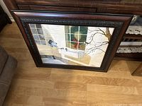 Framed oil painting showing exterior window scene with two birds, figure in red inside through window, and tree branch, resting on floor leaning against furniture.