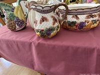 Four Fitz and Floyd Omnibus ceramic pieces displayed on a maroon tablecloth, showing decorative grape and fruit designs.