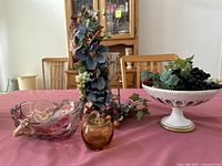 Photo showing the entire lot on a table with a pink tablecloth, including the footed fruit bowl with artificial grapes, pink-handled glass bowl, amber glass apple, and candle holder with floral decorations.