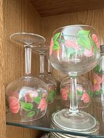 Group of four floral painted wine glasses with pink flowers and green leaves displayed in a wooden cabinet.