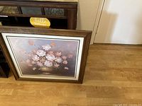 Framed floral print leaning against a wall on wood floor, showing full artwork and frame.